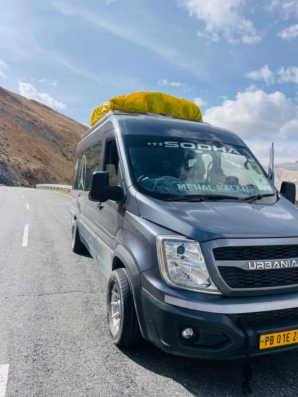 Grey SODHA-branded van with luggage on the roof, on a mountain road in Ladakh